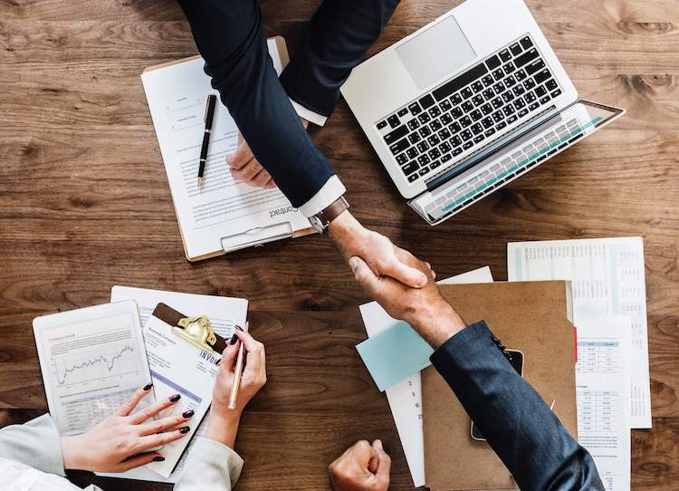 Project managers in business attire shake hands at a table with stacks of schedules, contracts, and invoices.