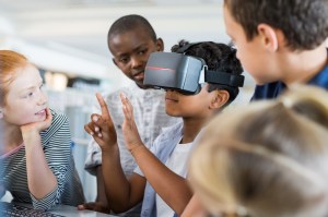 A group of students gathers around to observe their classmate wearing a pair of virtual reality goggles.