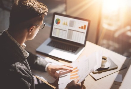 young male accountant looks at charts on a laptop screen and printed documents