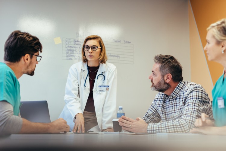 A hospital CEO stands at a conference table leading a meeting.