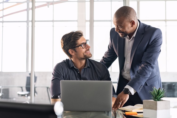 Two professionally dressed men, one standing and one seated at a laptop computer, exchange smiles.