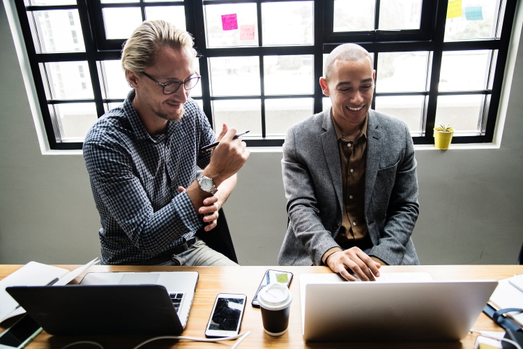 Two public relations professionals sit together at a table by a window looking at a laptop