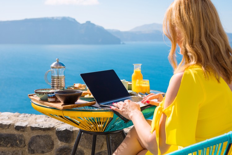 A travel writer is having breakfast on a lakeside terrace while writing an article on a laptop.