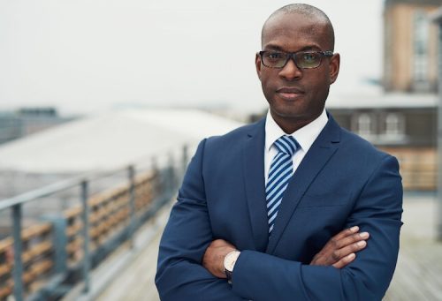 a male businessman wearing a suit stands outdoors with arms crossed