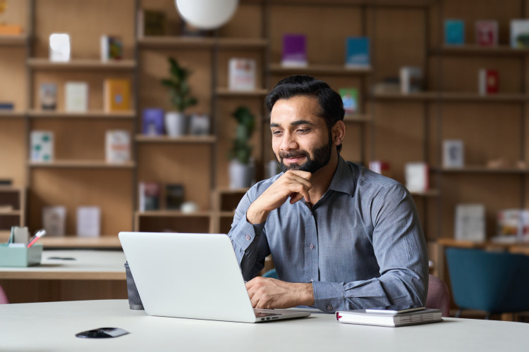 A freelance copywriter is jotting down ideas in a notebook while working on his laptop from home. 