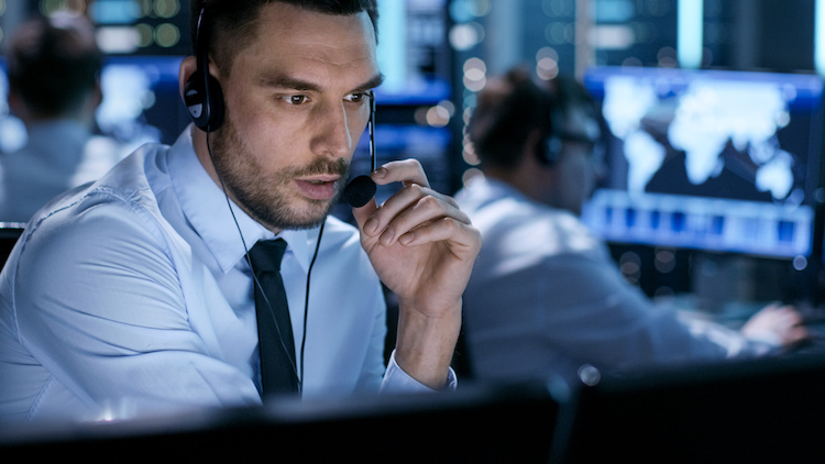 In Monitoring Room Technical Support Specialist Speaks into Headset. His Colleagues are Working in the Background.