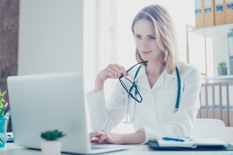 A health information manager works on a laptop at her desk while wearing a white coat