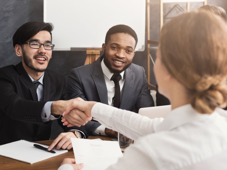 Two smiling men sit at a table across from a woman, and one of them shakes her hand.