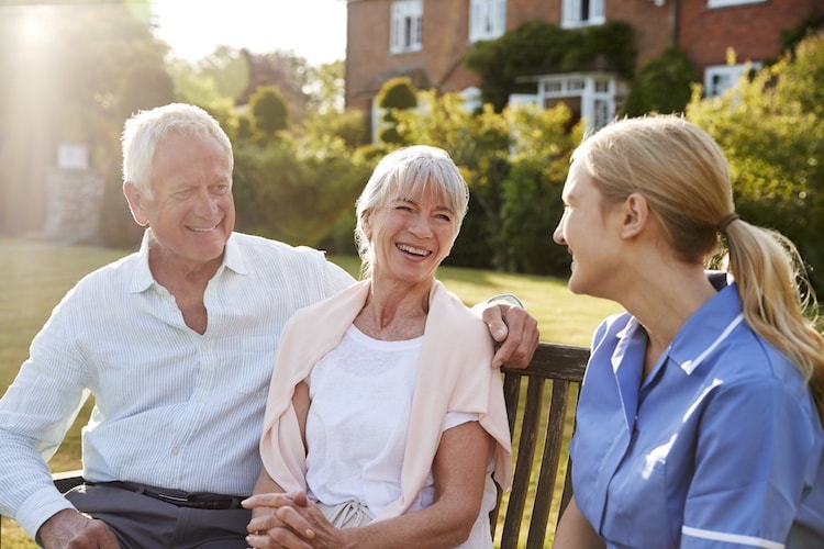 A retired elderly couple sit on a bench together at a retirement home.