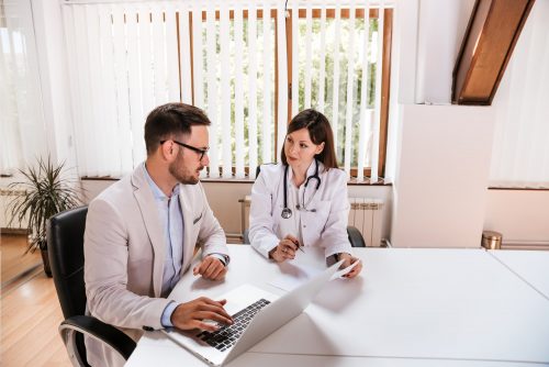 A man in a suit meets with a female doctor seated at a table in a conference room