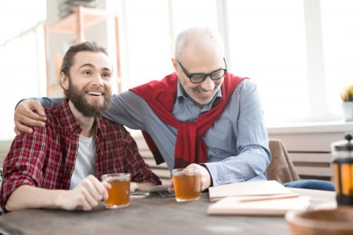 A younger worker and an older coworker laugh while drinking tea at a table with work documents on it.